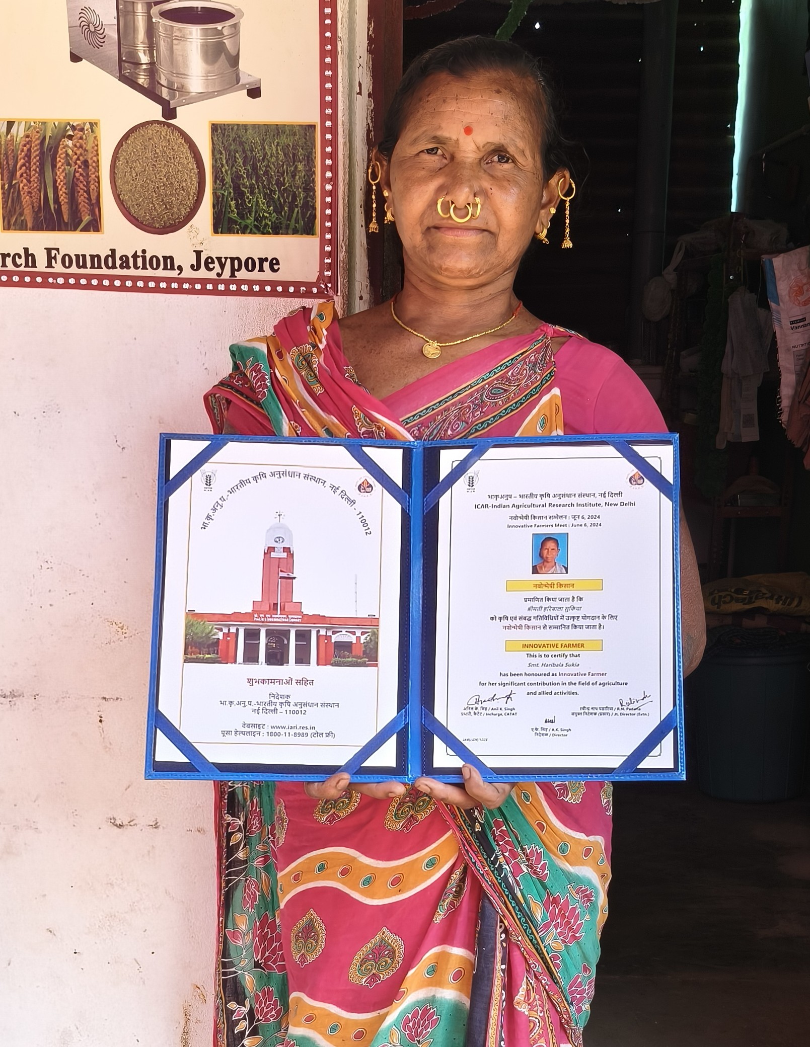 Horibola Sukia of Machara Village of Koraput District standing with her ICAR award that recognize Millet Seed Producer and Innovative Farmer. She is wearing a pink saree, is in her 60's and wears many brass earings and nose pericings.